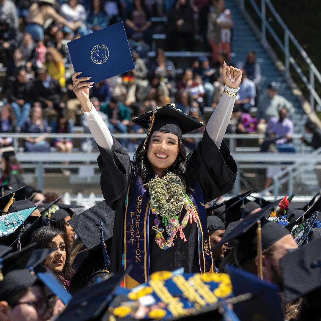 First Generation Graduate, standing with both her hands up in a sea of crowd holding her diploma