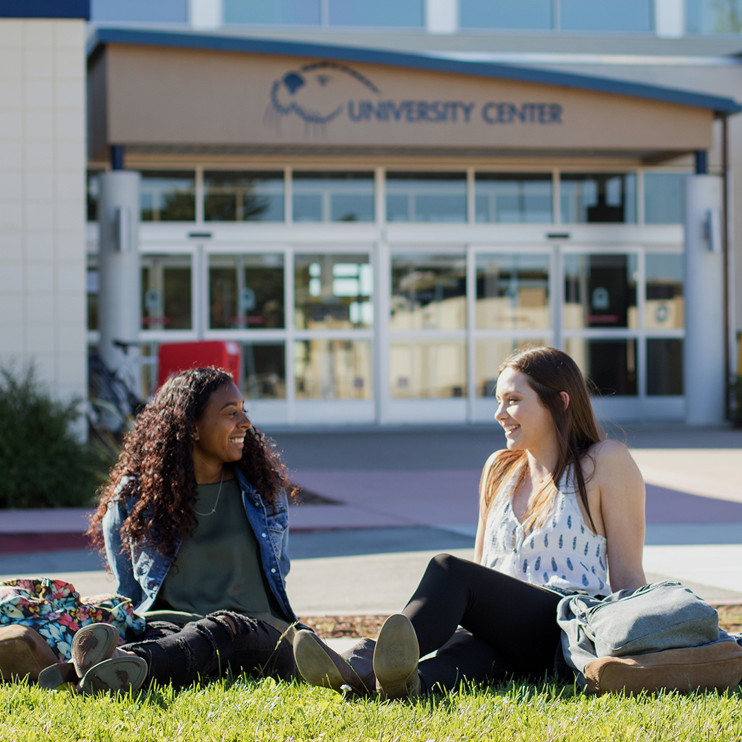 Two CSUMB students sitting at the lawn in front of the university center building