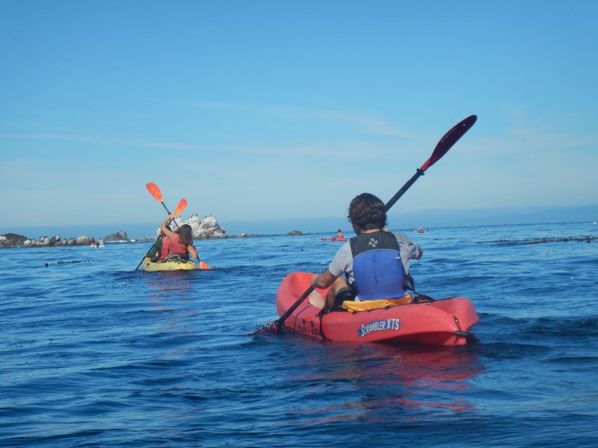 Three students in kayak in 2 kayaks on the ocean with rocks in the background.