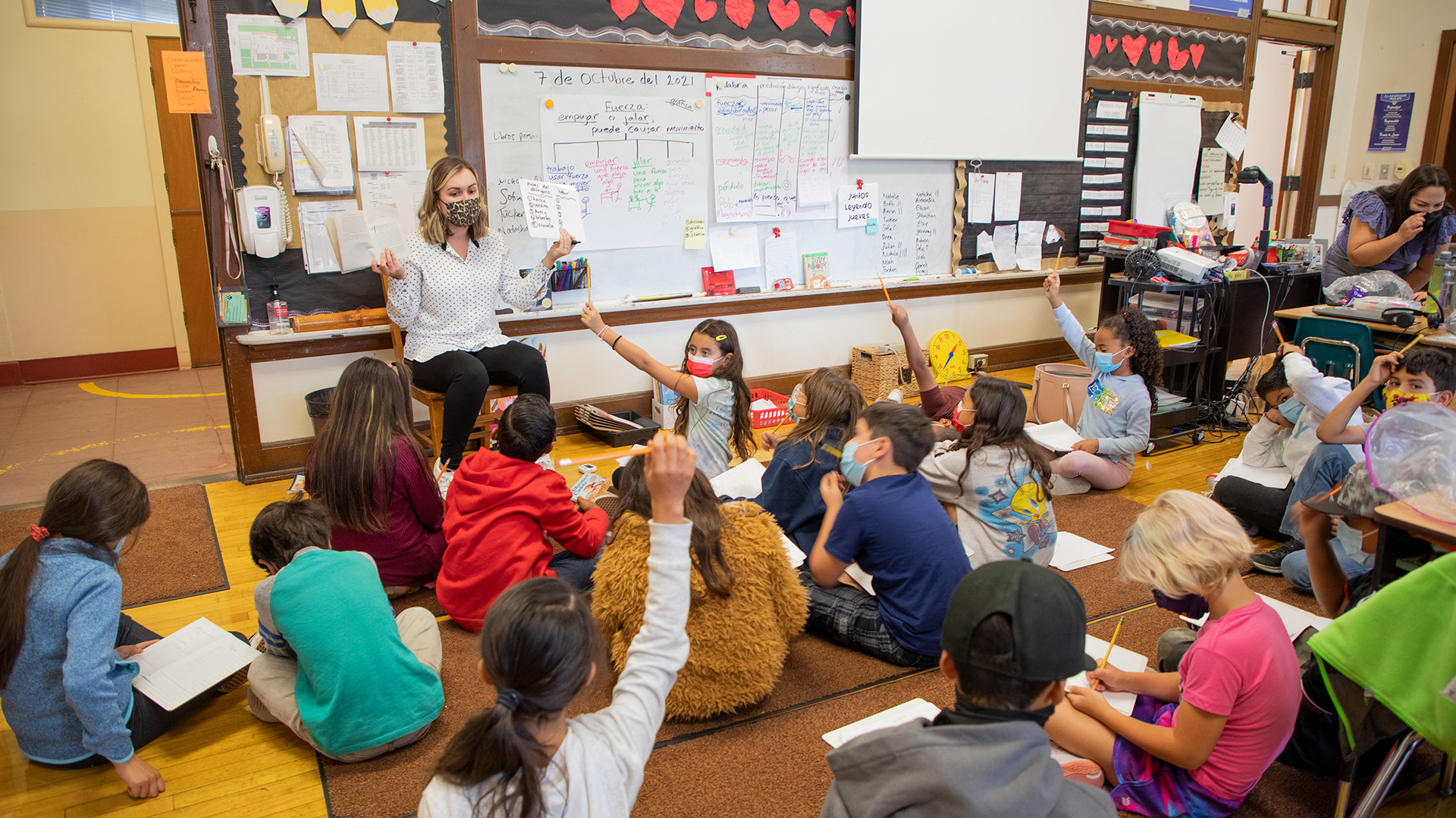 A teacher in a classroom and surrounded by kids pointing to a board