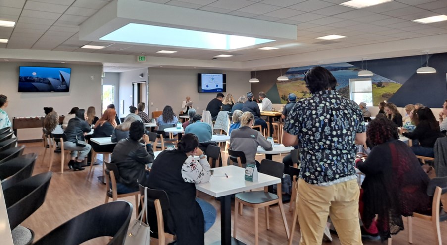 staff members listen to presentation on survey results, people standing, sitting, presentation on screen