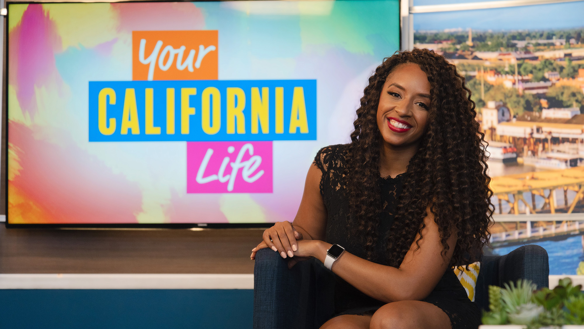 Desiree Sheppard sitting on a couch in front of a TV that shows a text graphic that reads 