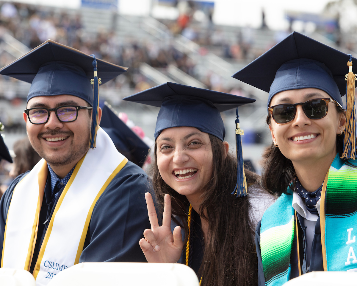 Three students sitting during their college convocation with one holding up a peace sign.